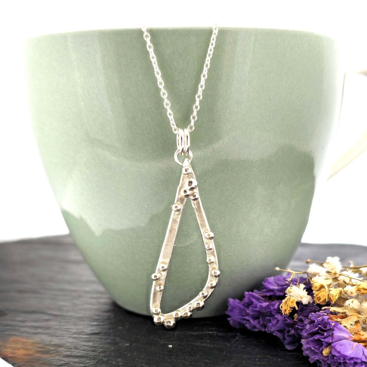 A silver asymmetrical pendant with a soft open triangle shape and silver balls, suspended from a silver chain. Shown on a green bowl with dried flowers.