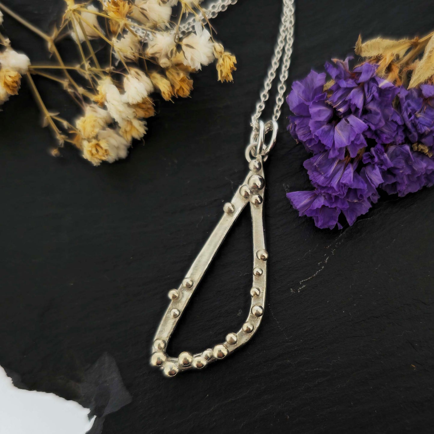 A silver asymmetrical pendant with a soft open triangle shape and silver balls, suspended from a silver chain. Shown on a black background with dried flowers.