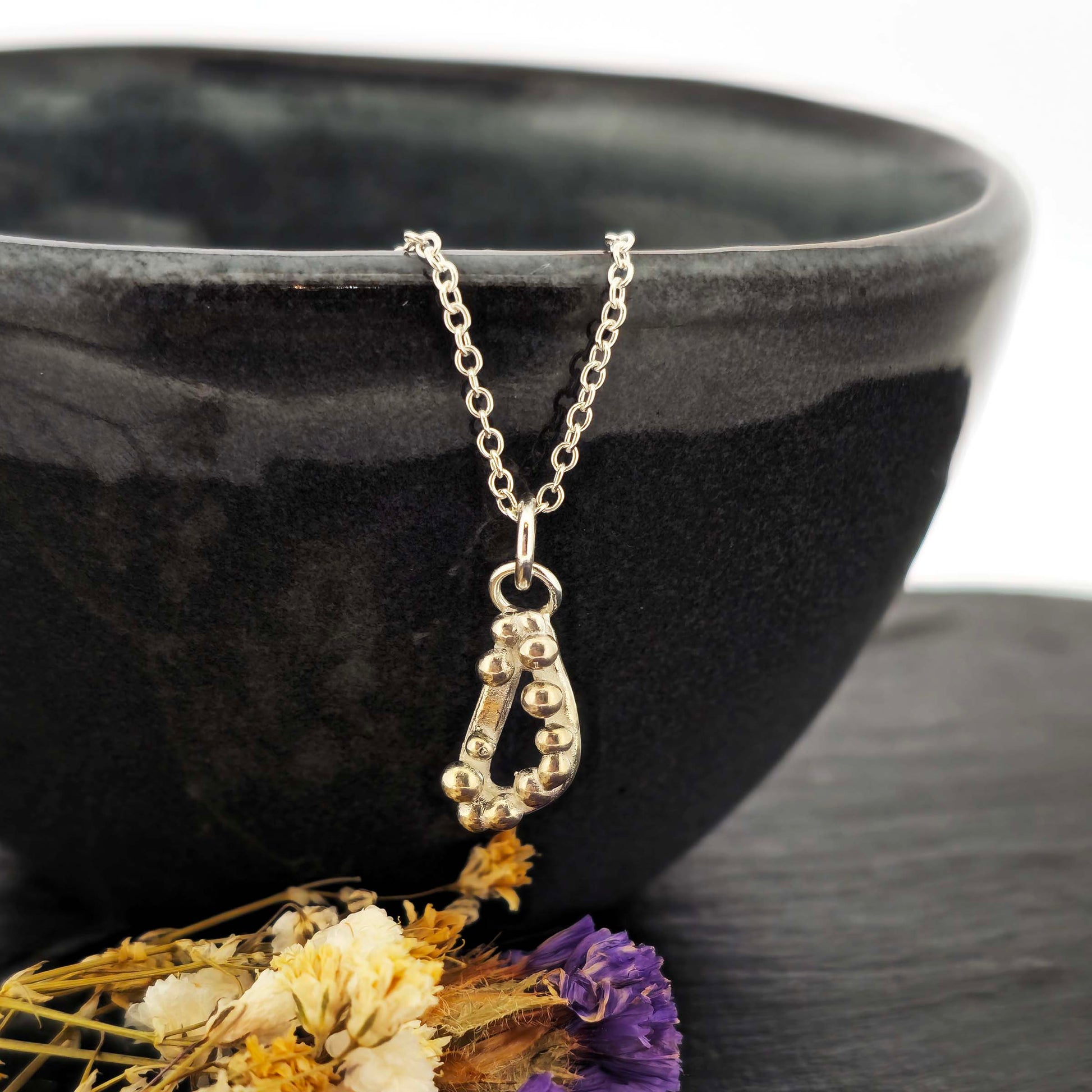 Silver asymmetric open triangle pendant with silver balls on a silver chain. Shown on a black bowl with flowers.