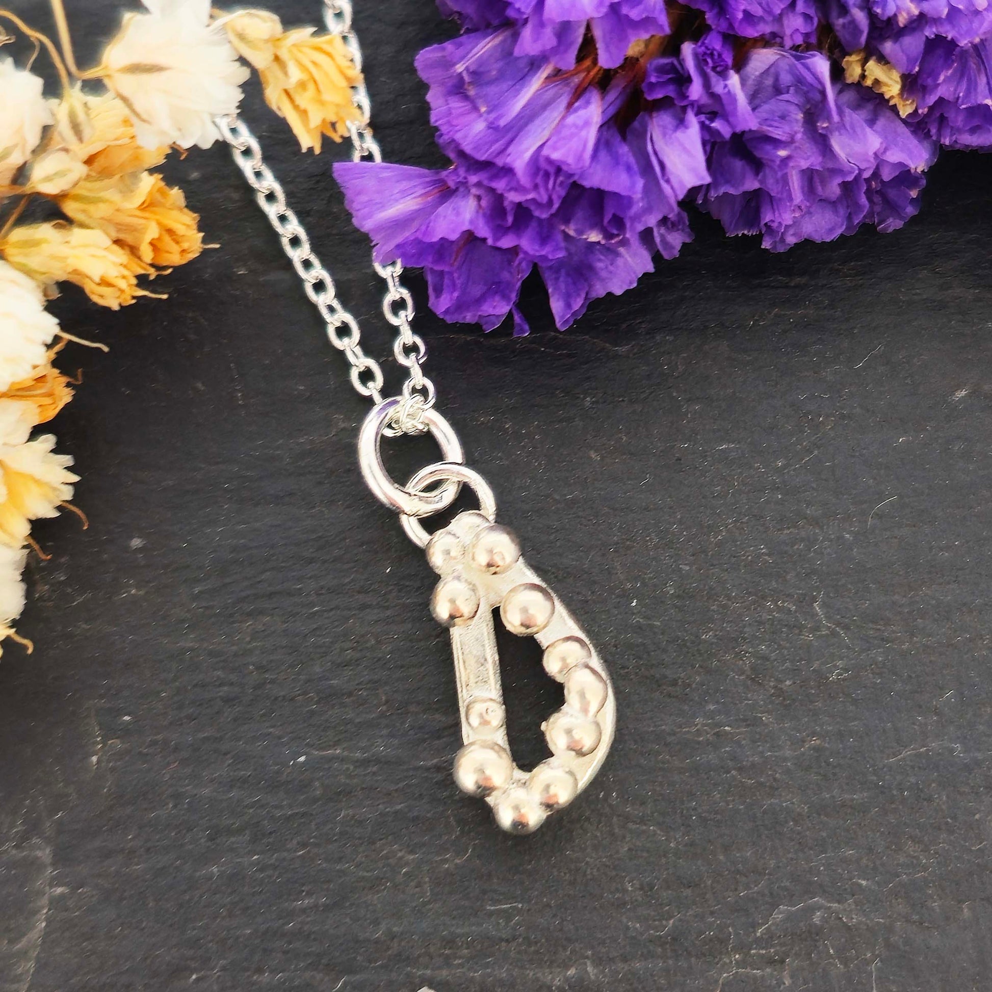 Silver asymmetric open triangle pendant with silver balls on a silver chain. Shown on a black background with dried flowers