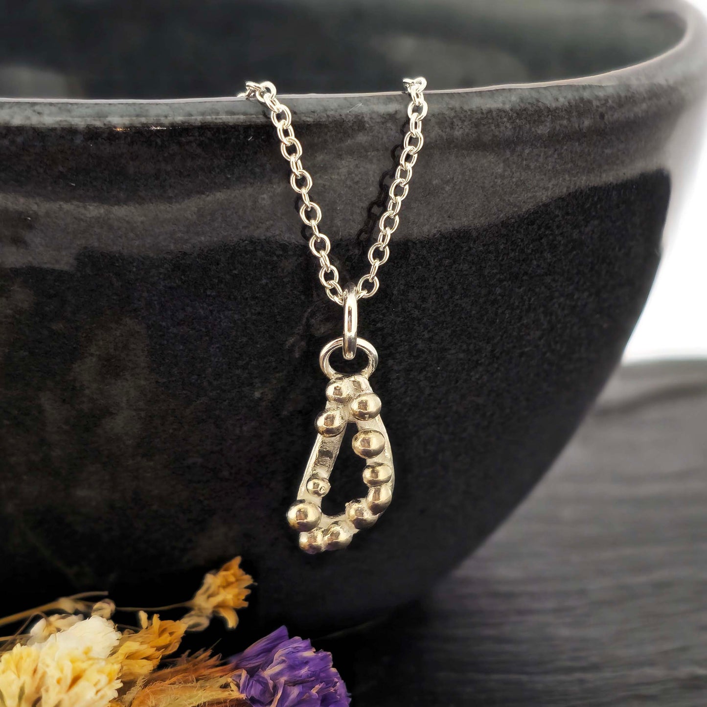 Silver asymmetric open triangle pendant with silver balls on a silver chain. Shown on a black bowl with flowers.