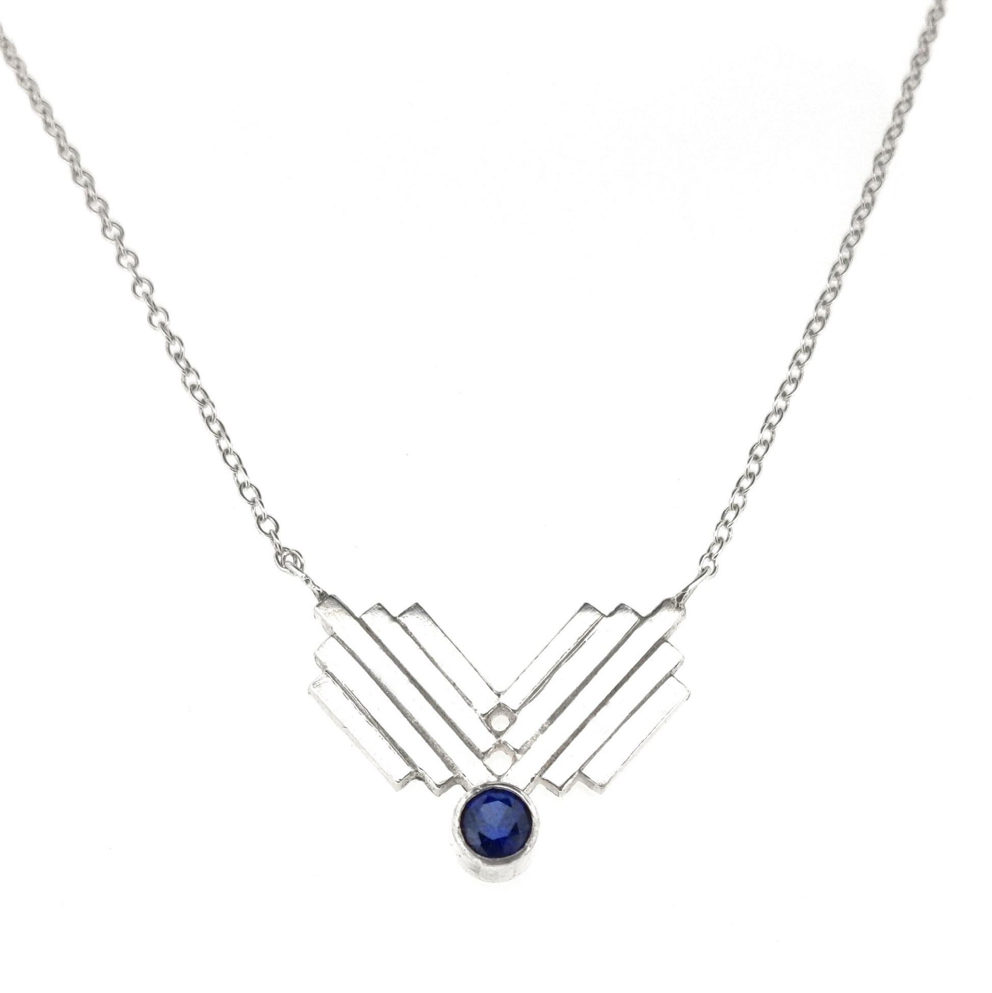 A silver Art Deco style necklace with a focal point of 2 lined elements and a dark blue sapphire gemstone.