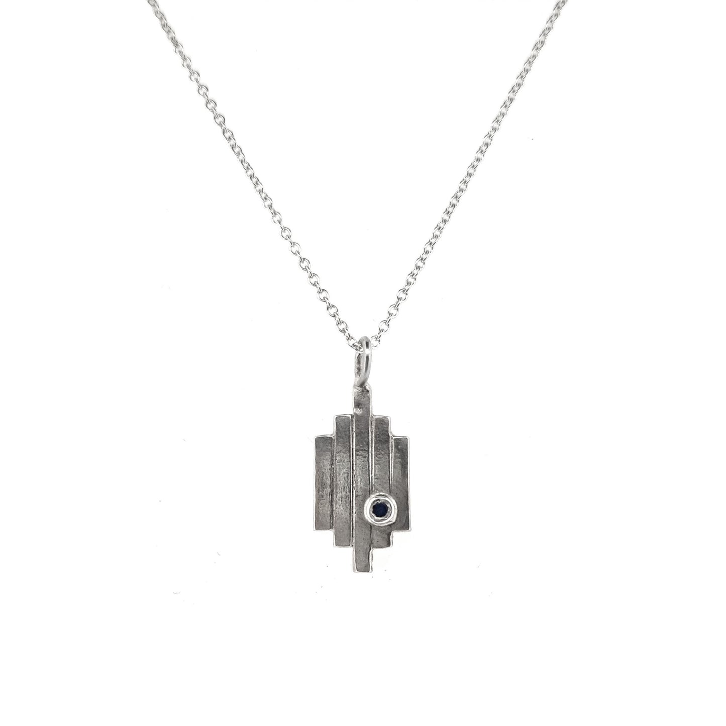 A silver Art Deco style pendant with 5 lines and a dark blue sapphire set off-centre. On a silver chain.