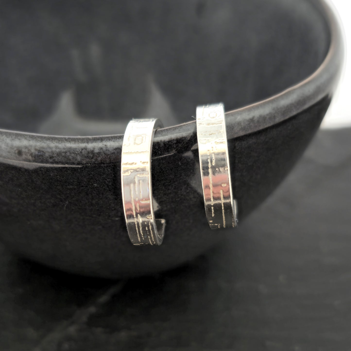 A pair of silver flat hoop earrings with a square and line pattern on. Pictured on the edge of a black bowl.