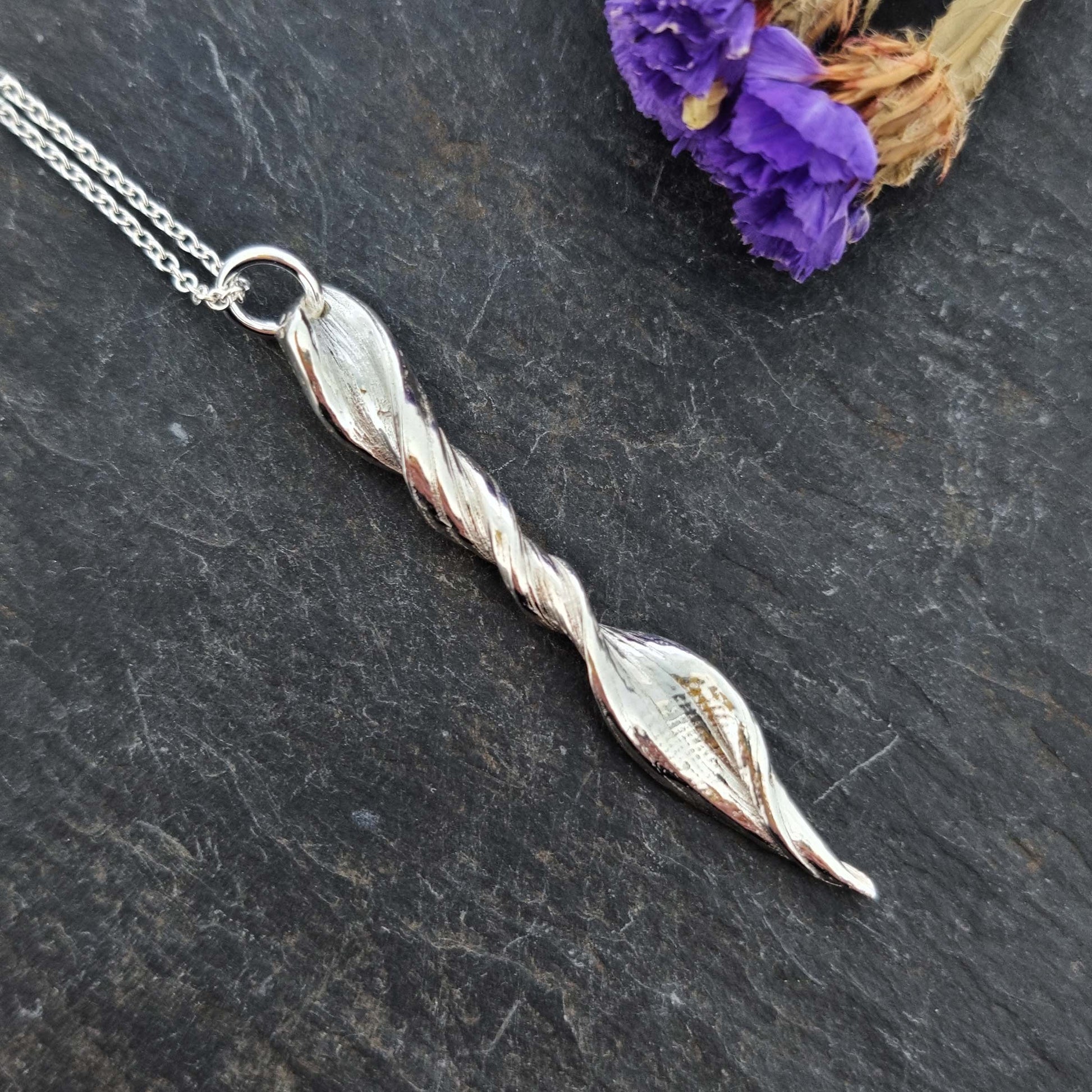 A silver twist pendant with line pattern on a silver chain. Pictured with flowers.