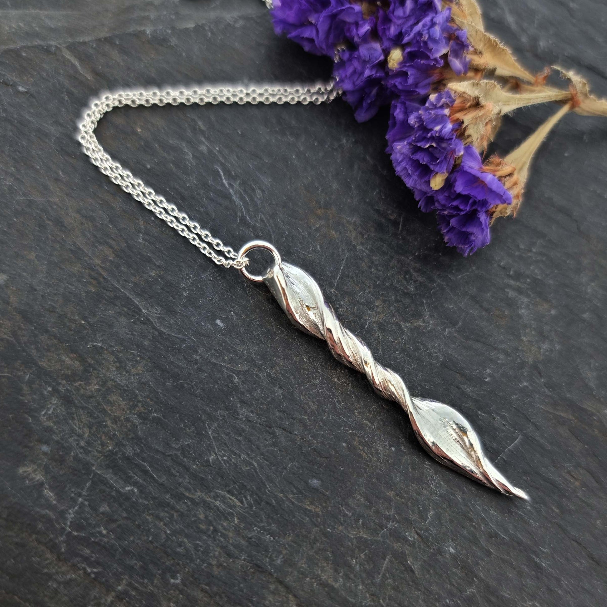 A silver twist pendant with line pattern on a silver chain. Pictured with flowers.