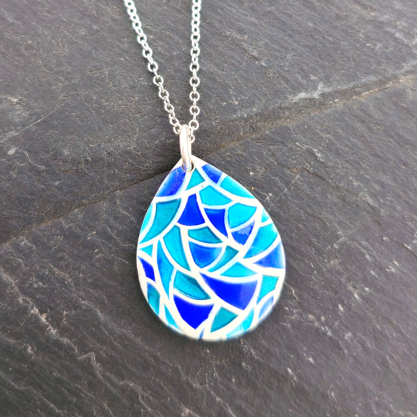 A teardrop shaped silver pendant with geometric cells of blue, green and turquoise enamel. On a silver chain. Pictured on a black background. Handmade. Mijoux Jewellery.