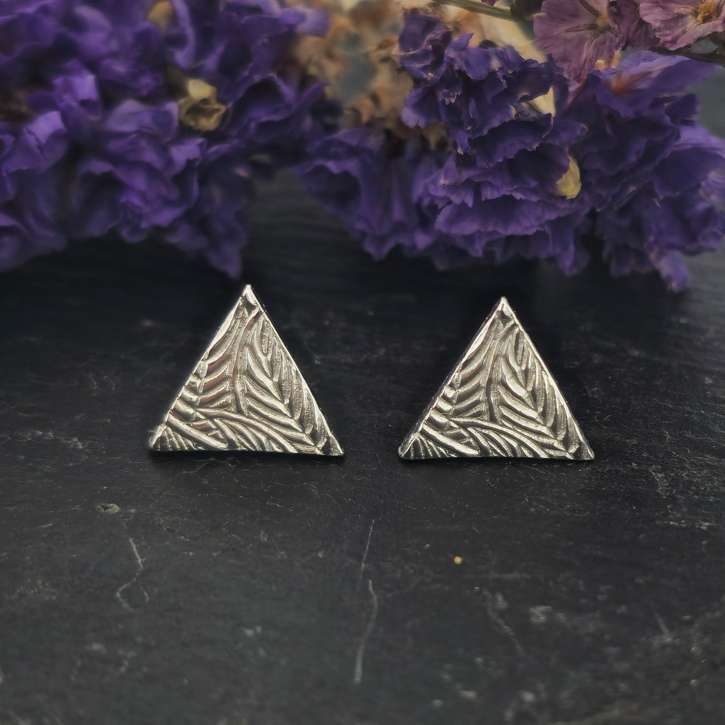 Sterling silver triangle stud earrings with a line leaf pattern. Shown on slate with flowers.