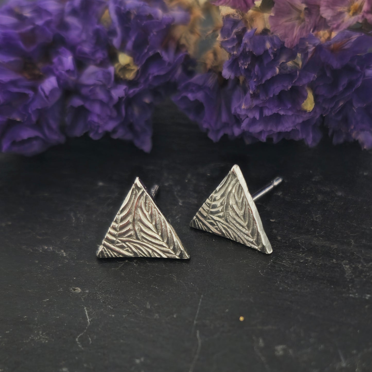 Sterling silver triangle stud earrings with a line leaf pattern. Shown on slate with flowers.