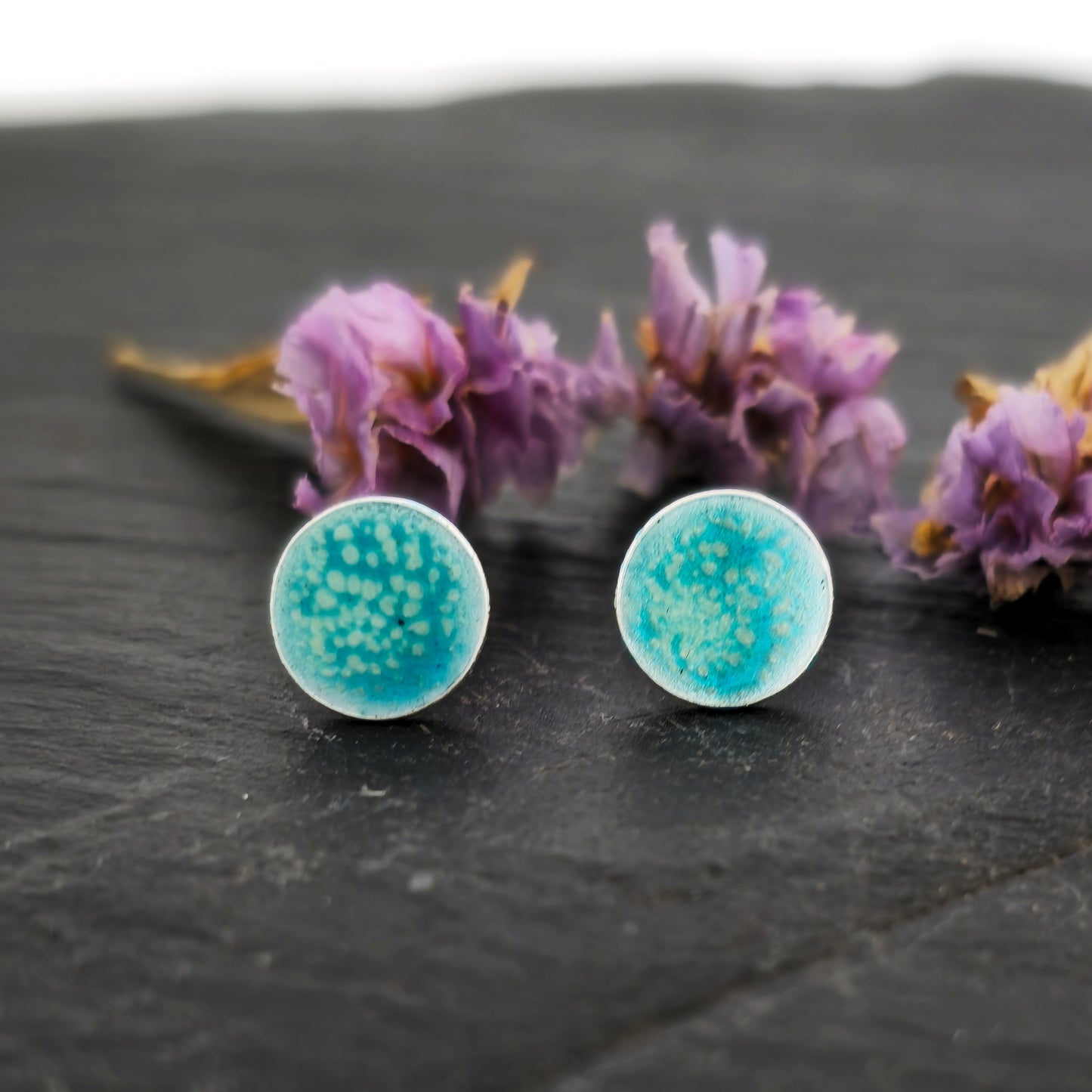 Round silver stud earrings with a turquoise and clear speckled enamel. Shown on slate with flowers.