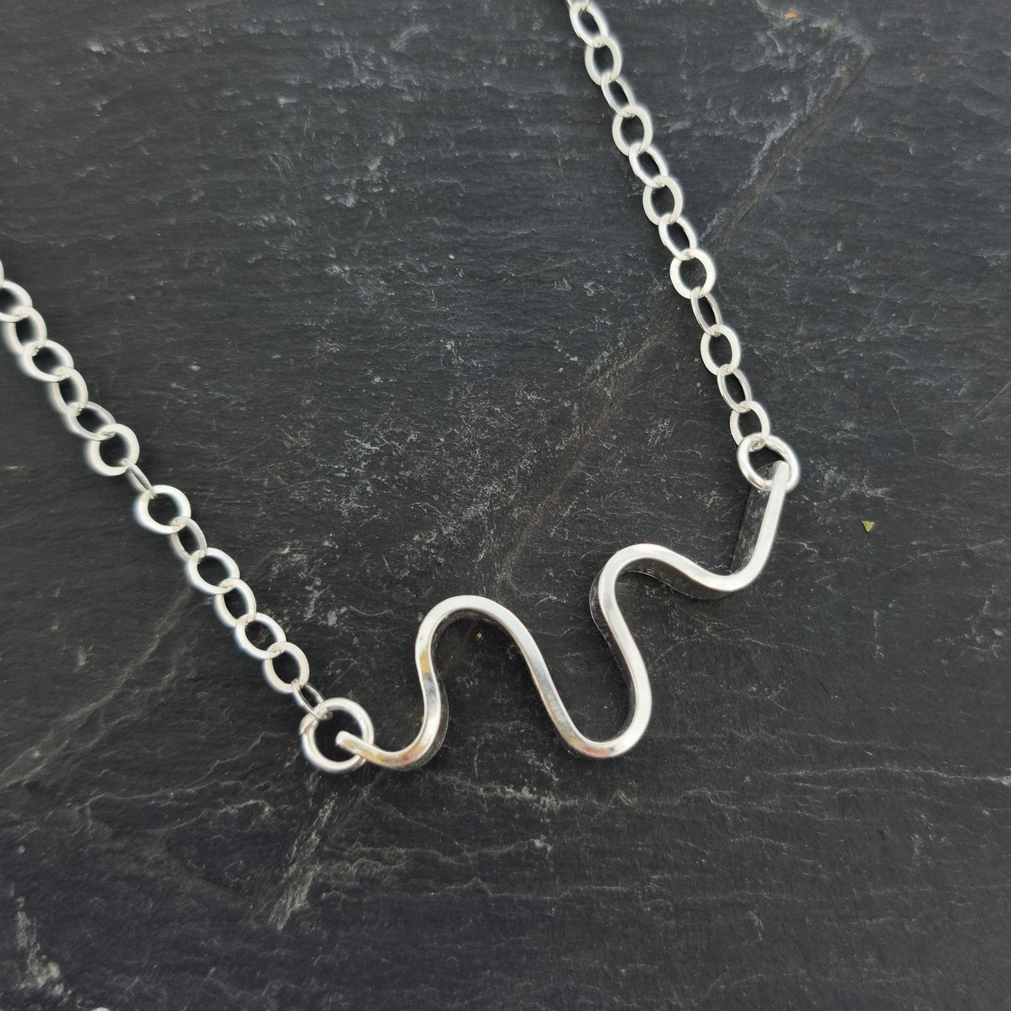 A silver necklace with an open chain and a wave of silver at the center. Shown on slate.