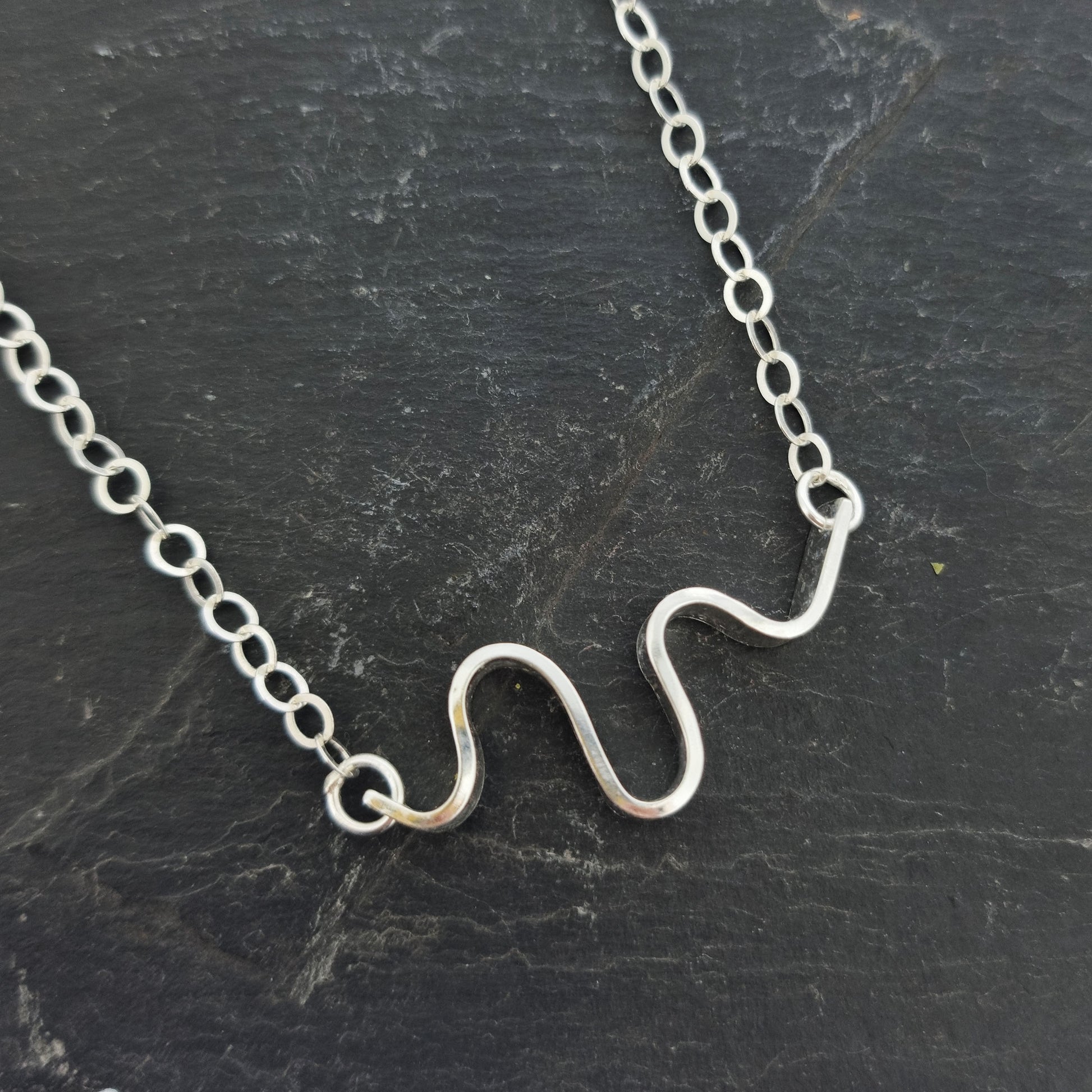 A silver necklace with an open chain and a wave of silver at the center. Shown on slate.
