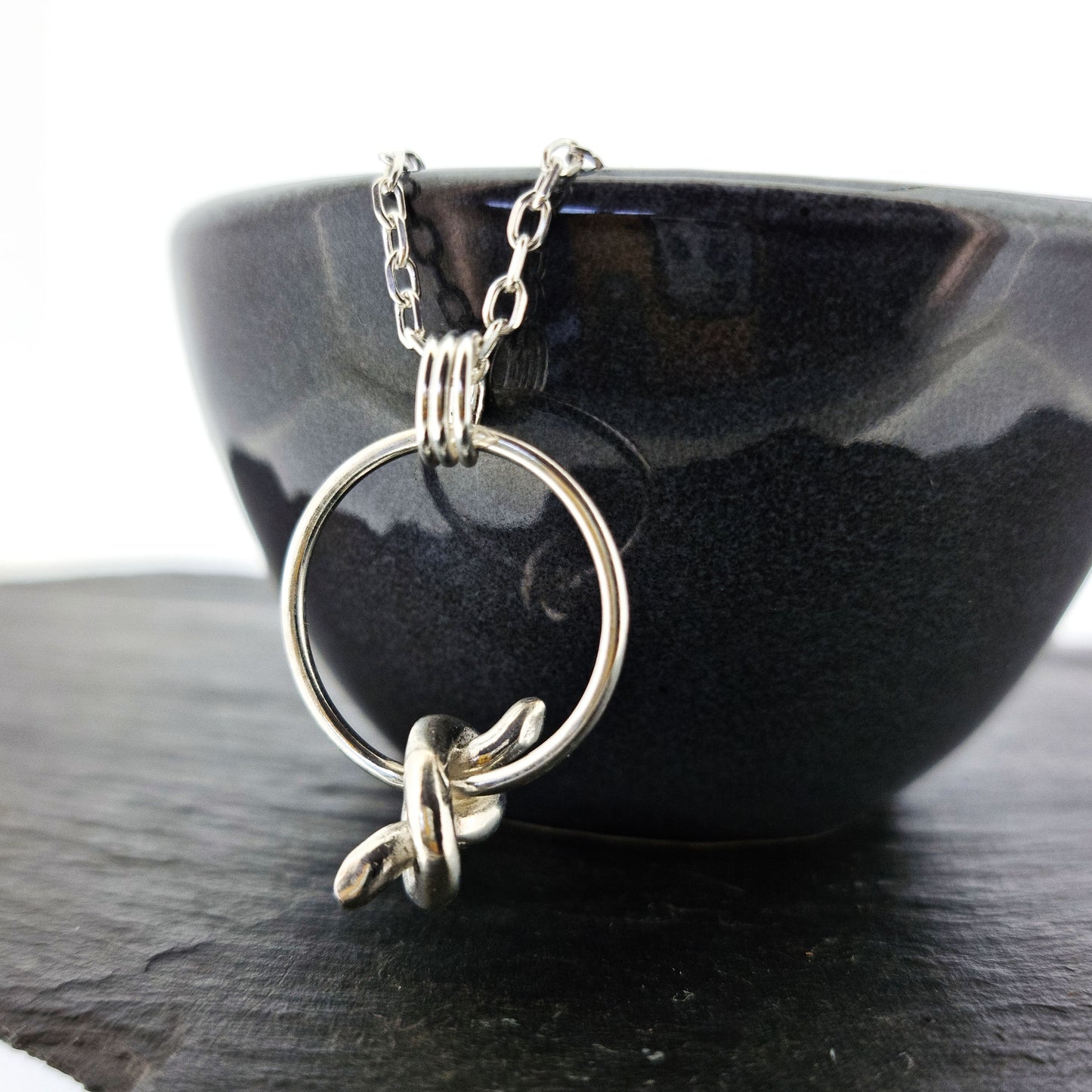 Silver circle pendant with spinning knot charm and triple circle bail. Suspended on a silver chain. Pictured on a black bowl.