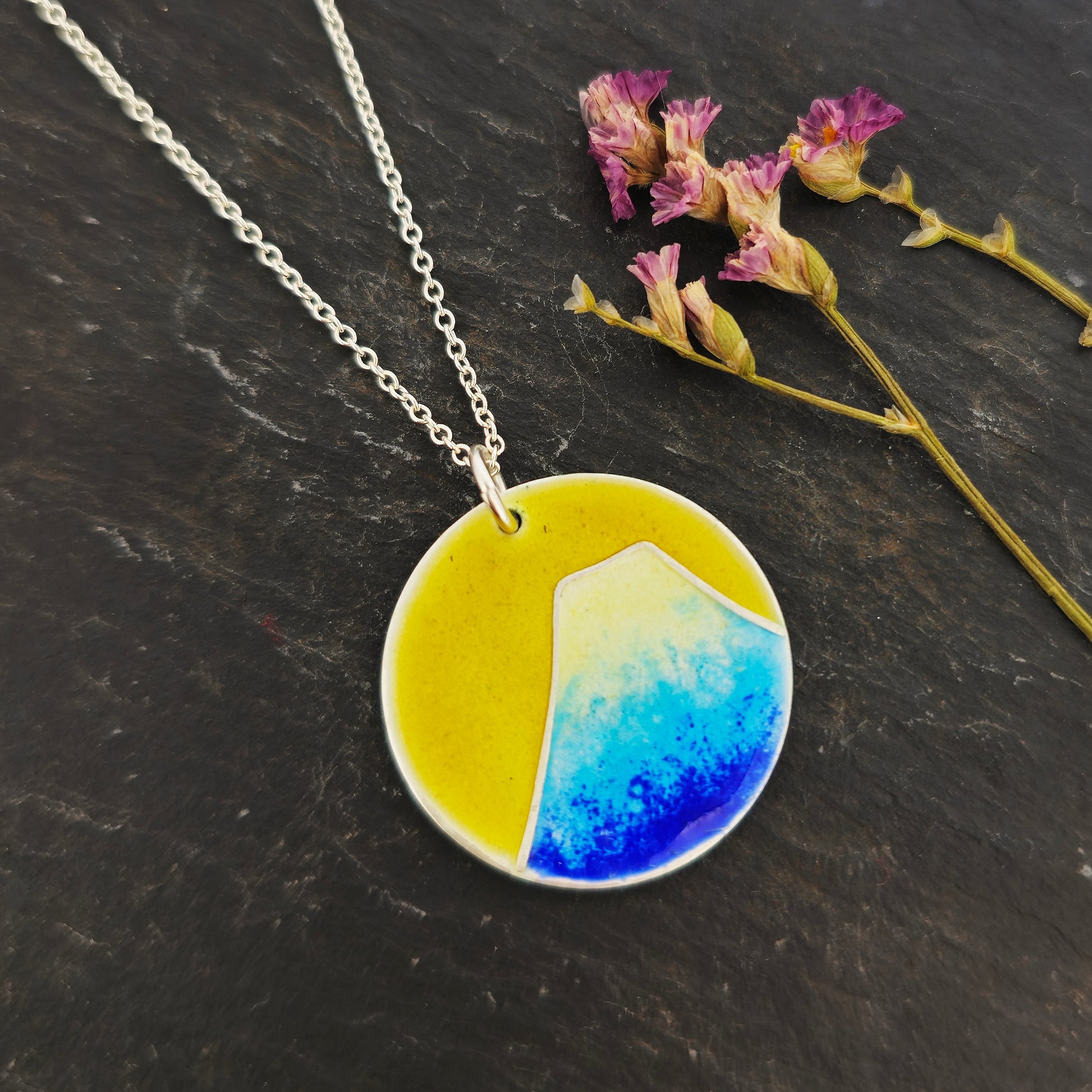 Round silver pendant with a mountain on in graduated blue enamel and a white top and a yellow enamel background. Suspended from a silver chain. Shown on slate with flowers.