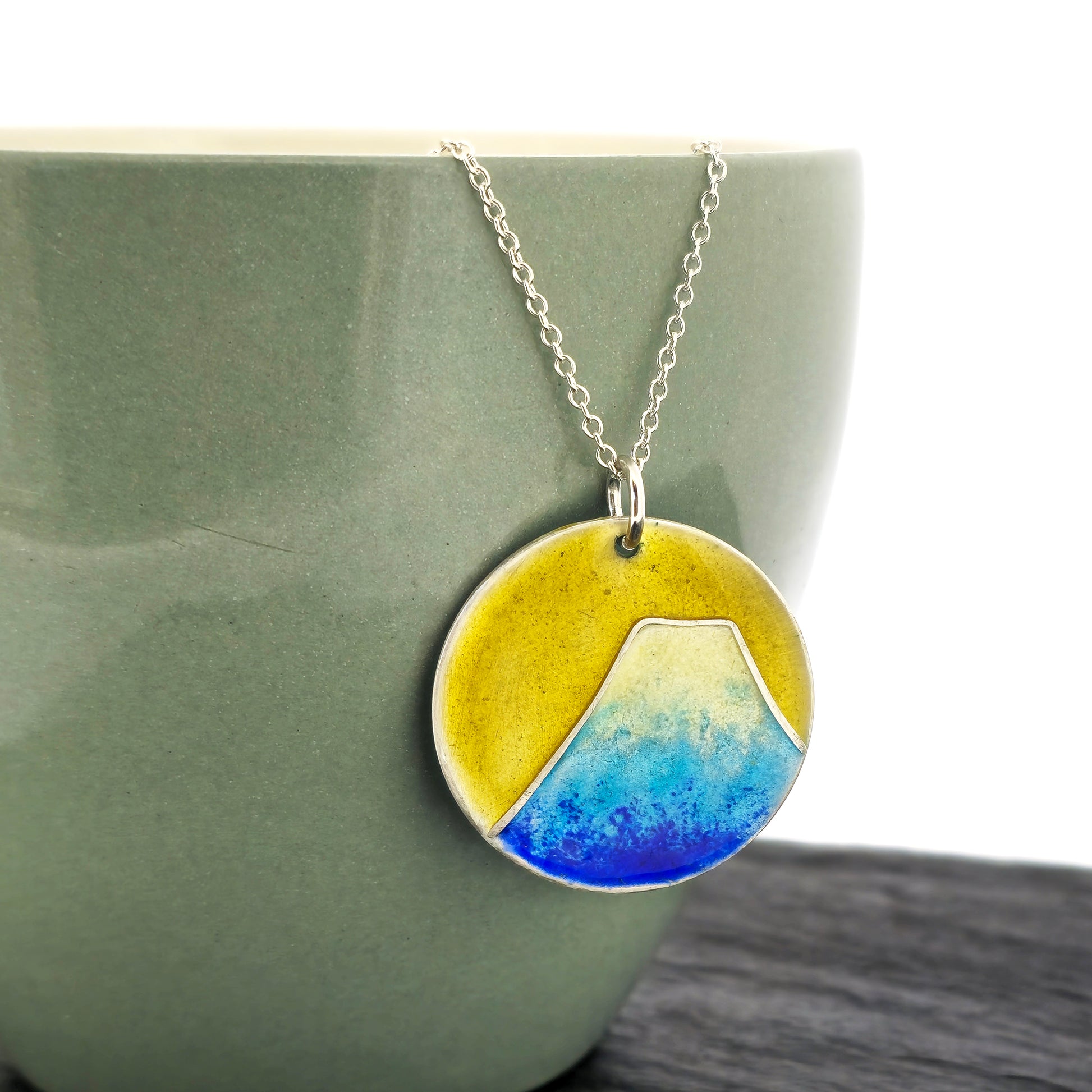 Round silver pendant with a mountain on in graduated blue enamel and a white top and a yellow enamel background. Suspended from a silver chain. Shown on a cup.