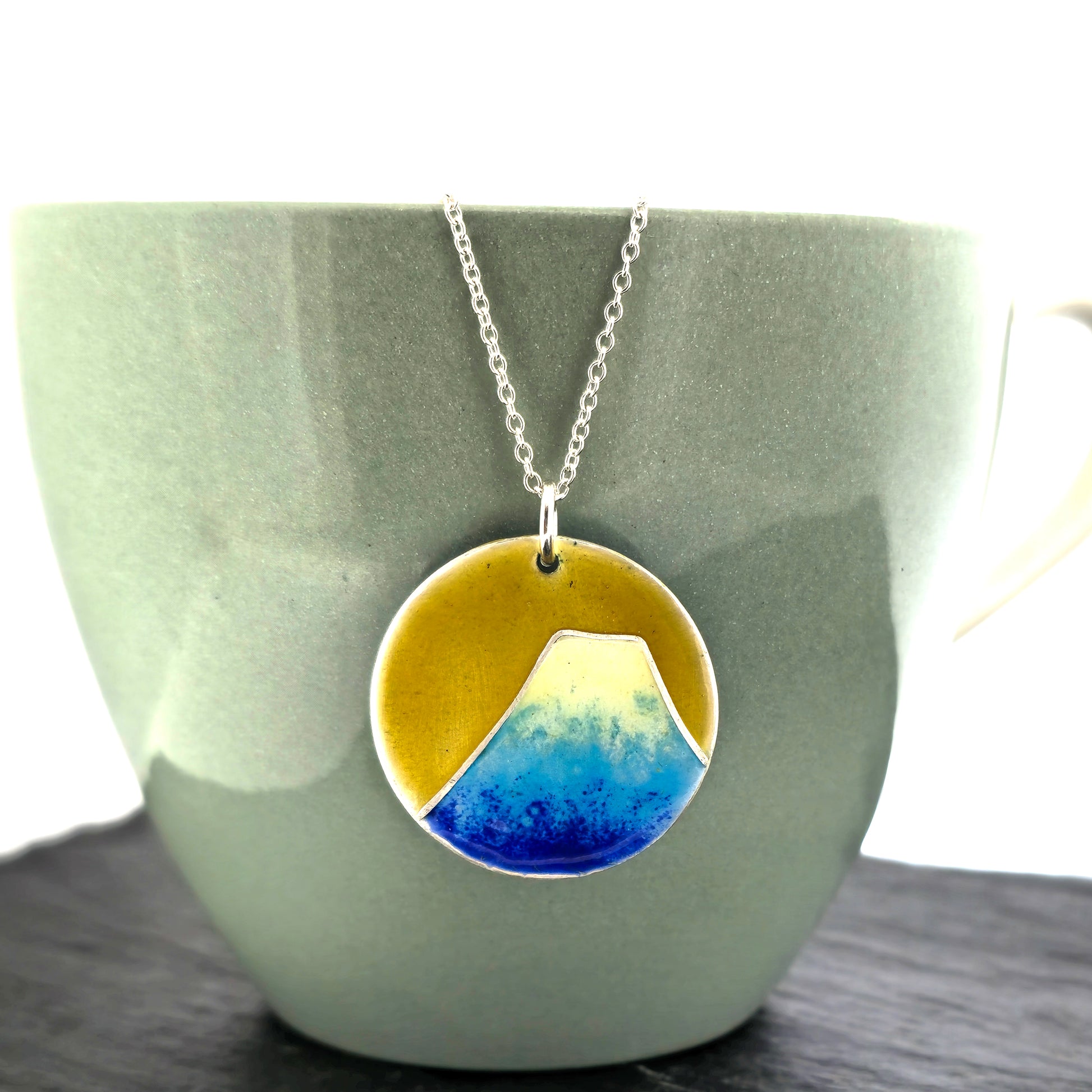 Round silver pendant with a mountain on in graduated blue enamel and a white top and a yellow enamel background. Suspended from a silver chain. Shown on a cup.