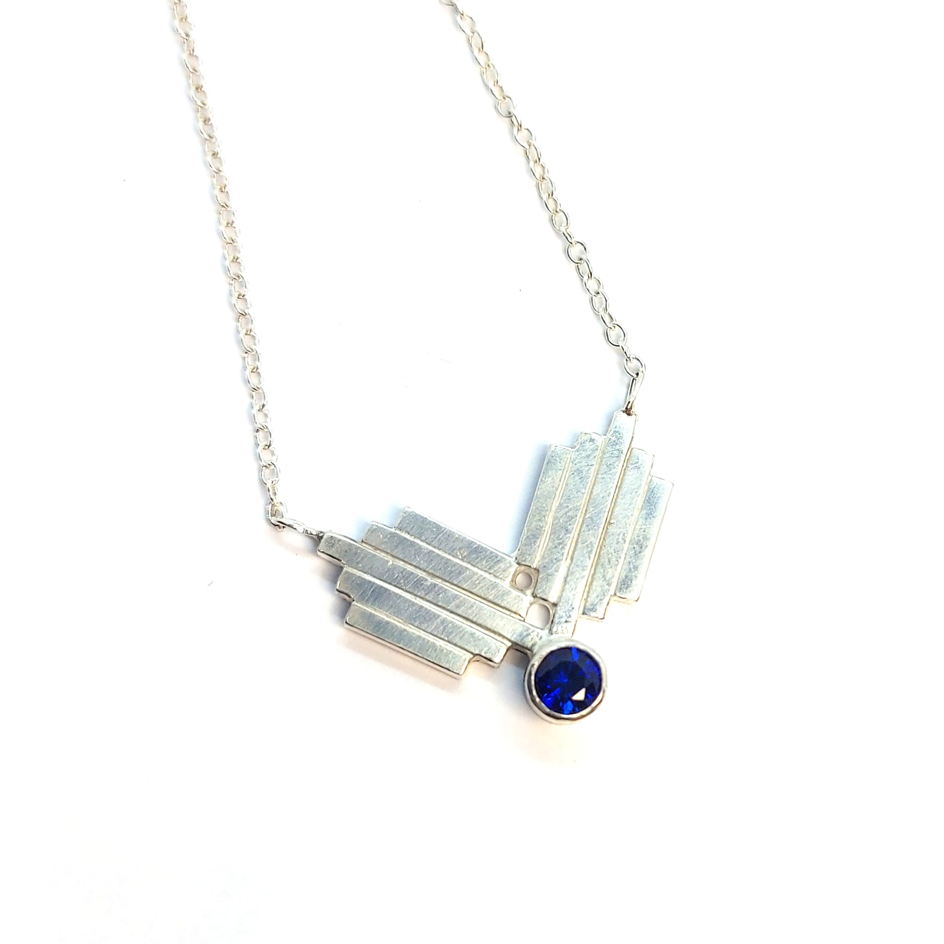 A silver Art Deco style necklace with a focal point of 2 lined elements and a dark blue sapphire gemstone.