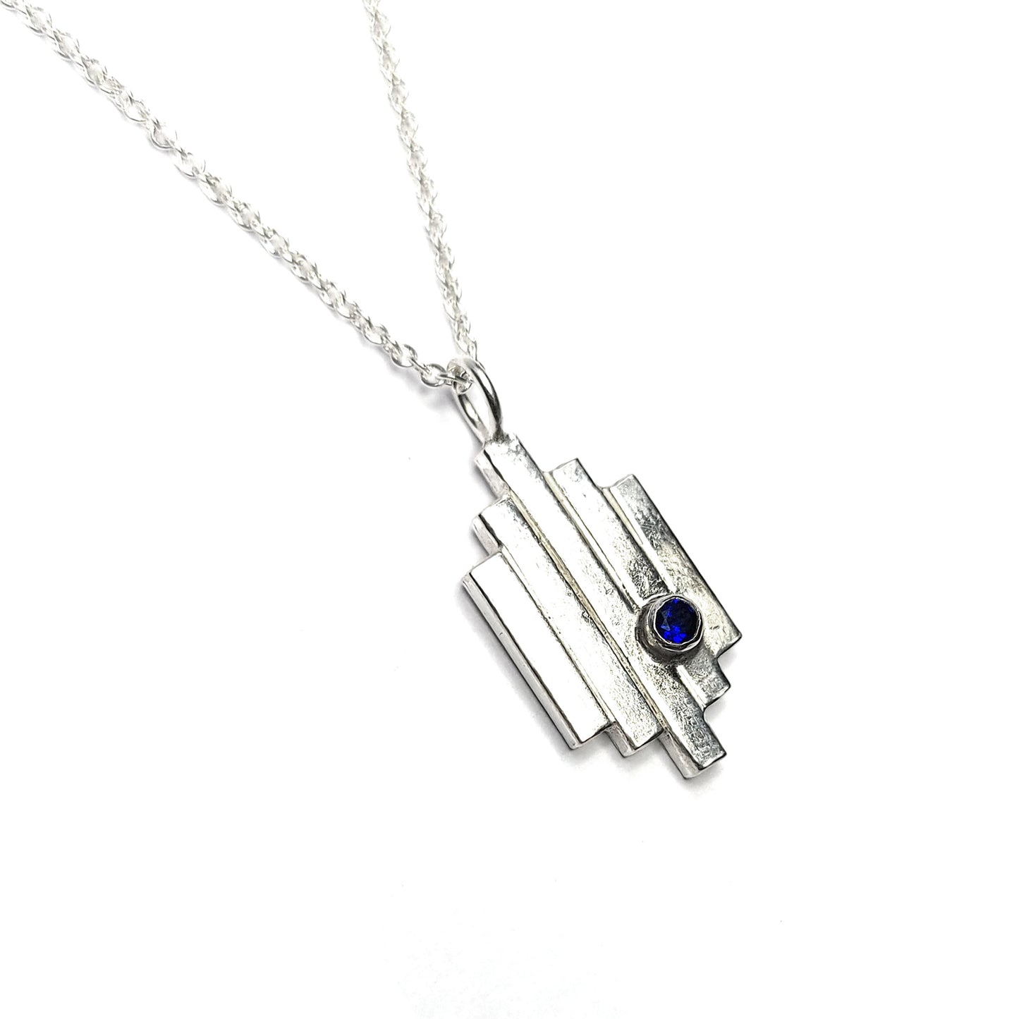 A silver Art Deco style pendant with 5 lines and a dark blue sapphire set off-centre. On a silver chain.