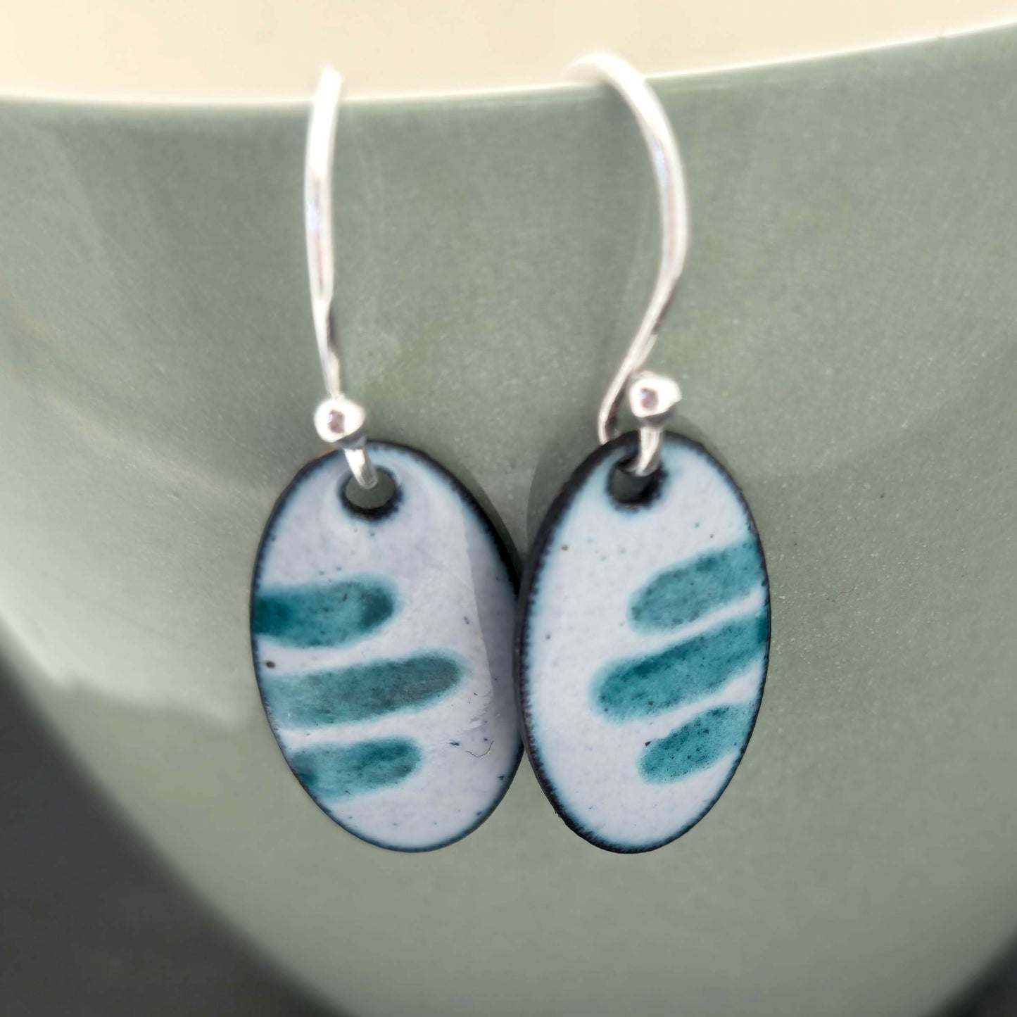 Oval-shaped enamel earrings with teal and light grey pattern on a green ceramic surface