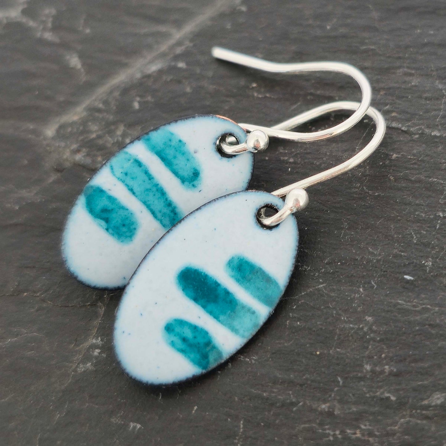 Pair of oval enamel earrings with teal stripe pattern on a light grey background with silver ear hooks on a slate background