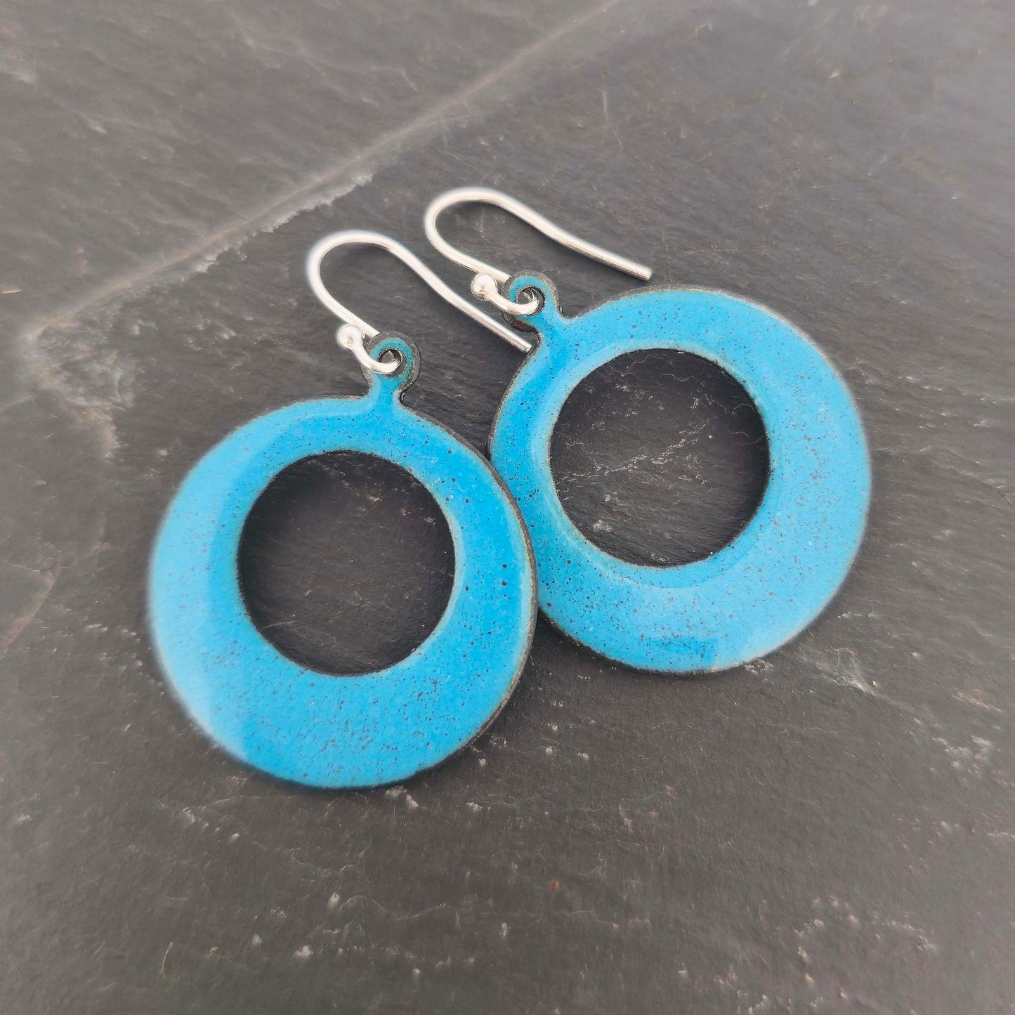 Enamel drop earrings featuring an open circle with teal blue enamel and a dusting of red enamel. On silver ear hooks. Shown on slate.