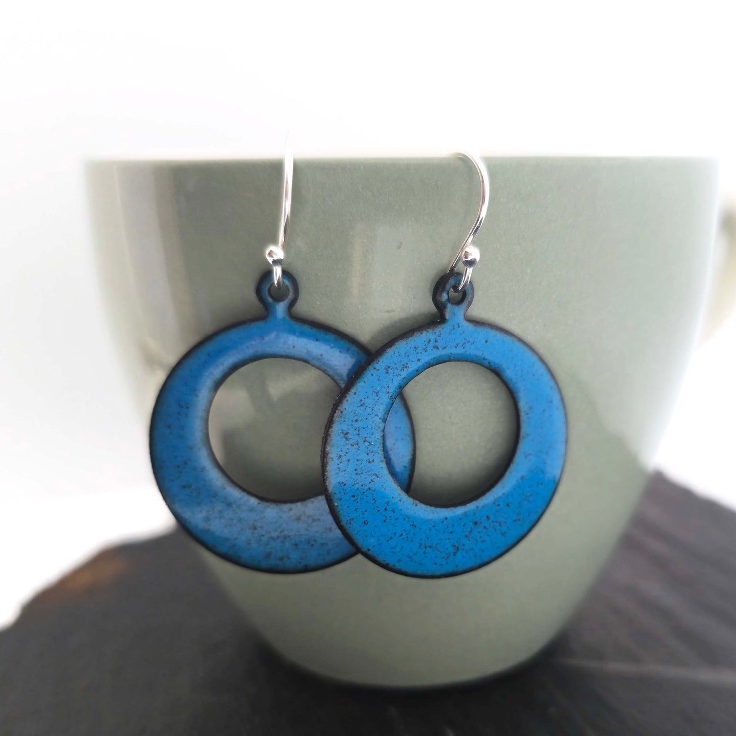 Enamel drop earrings featuring an open circle with teal enamel and a dusting of red enamel. On silver ear hooks. Shown hanging on a cup.