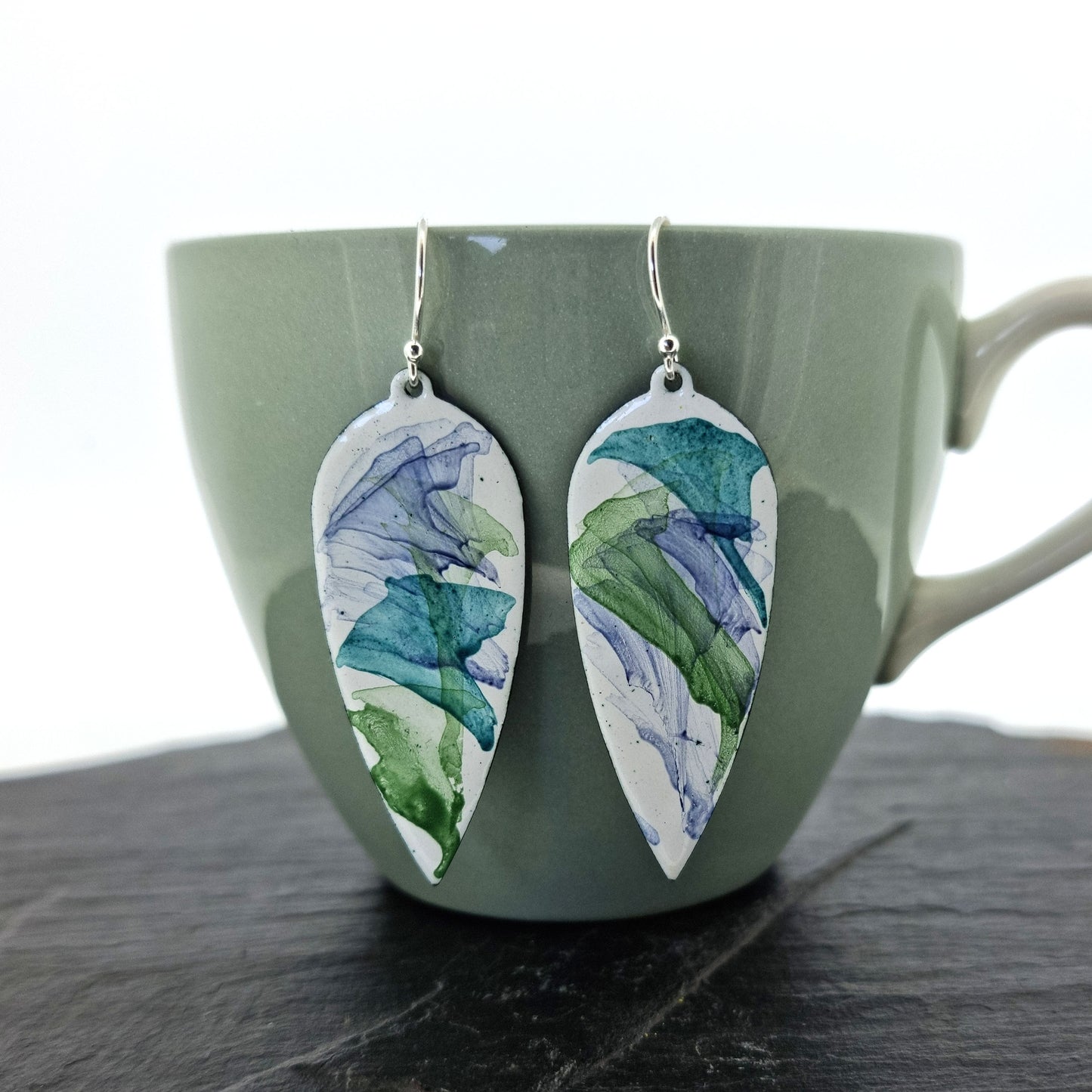 Large teardrop-shaped enamel drop earrings with splashes of dark blue and green on a white background. Pictured on a cup.