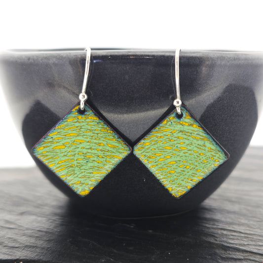 Diamond shaped drop earrings features a yellow and green scatter line enamel pattern. Suspended from silver ear hooks. Shown on a black bowl.
