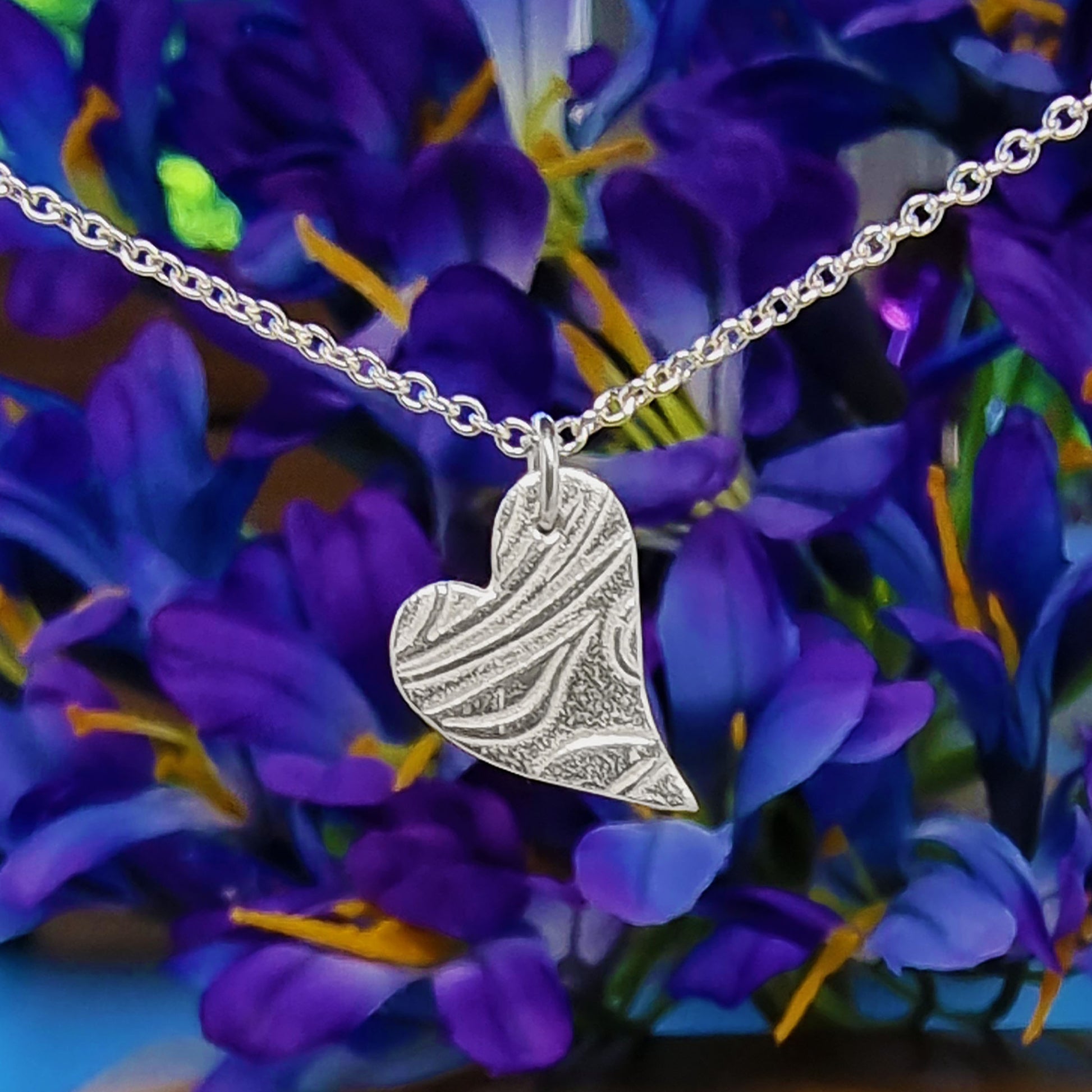 A silver asymmetrical heart pendant with a leaf and vine pattern suspended on a silver chain - small pictured with a floral background.