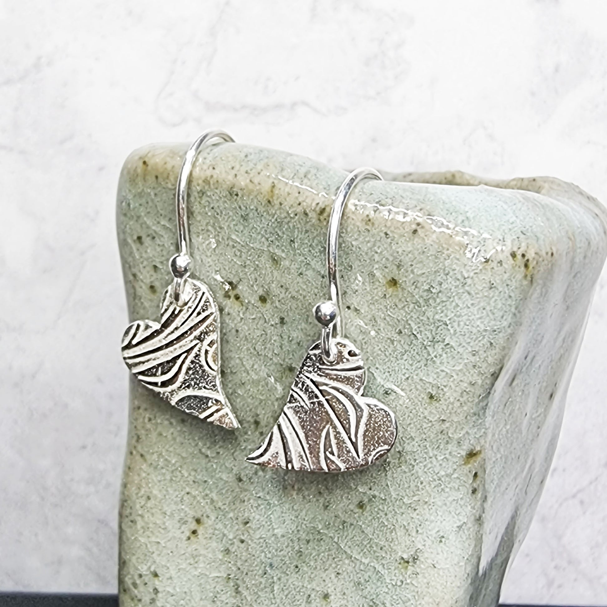 Silver heart drop earrings featuring an asymmetrical heart with a leaf and vine pattern suspended from silver ear hooks. Shown on a vase.