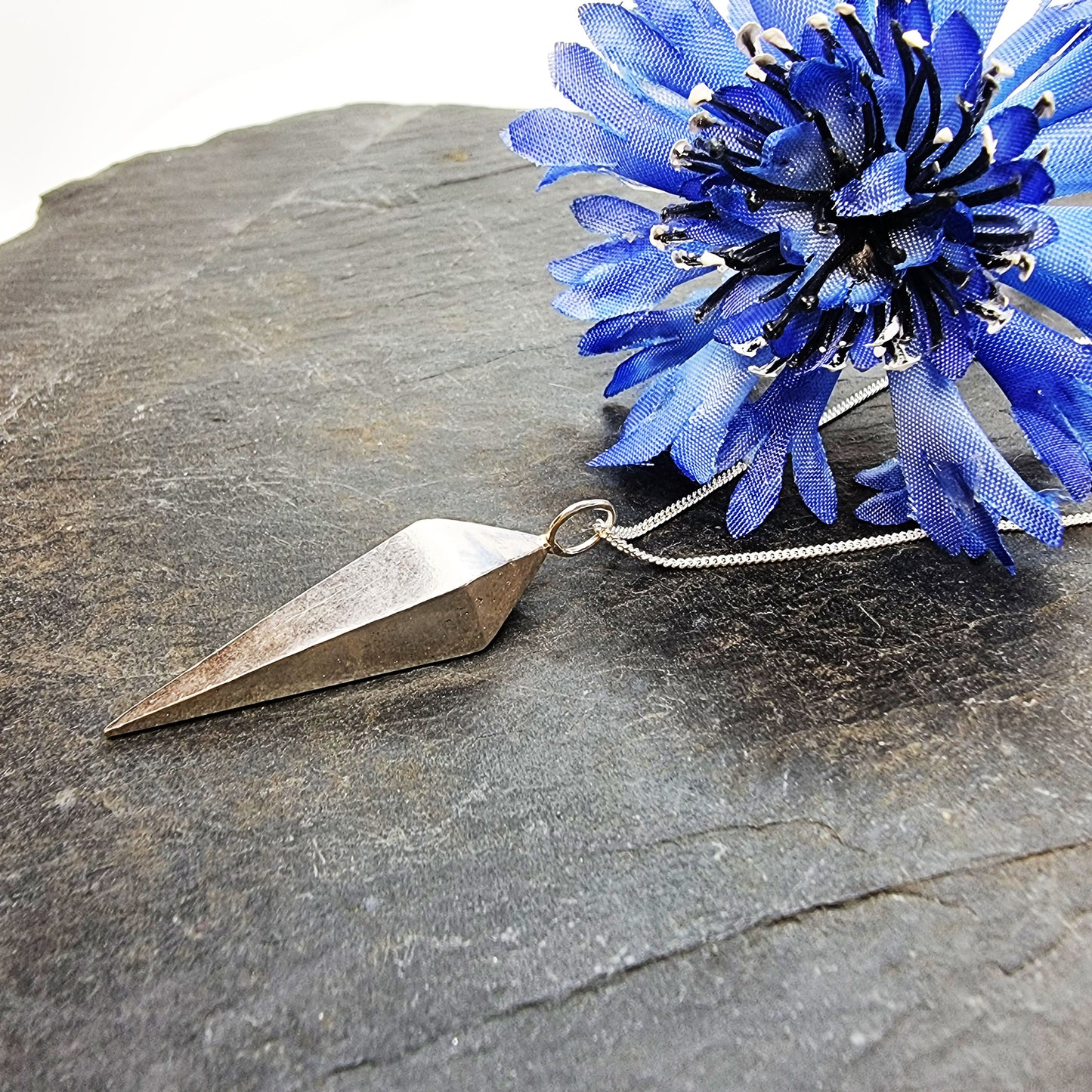 A silver geometric diamond shaped pendant on a silver chain. Pictured with a blue flower.