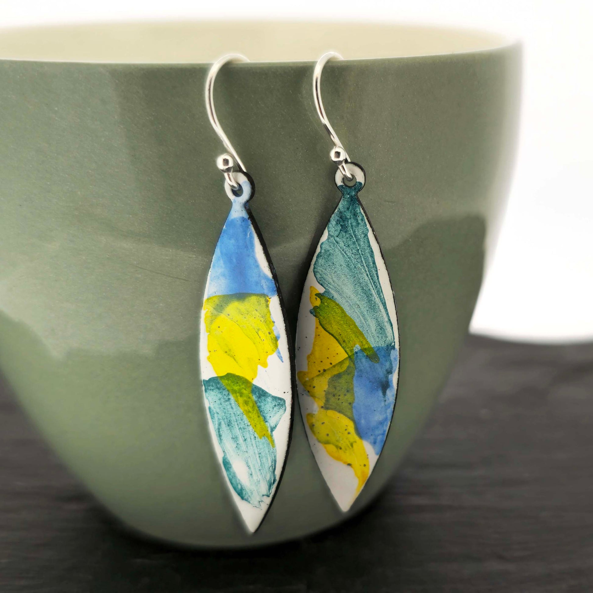 Navette shaped enamel drop earrings with green, blue and yellow splashes on a white background with silver ear hooks. Pictured on a cup.