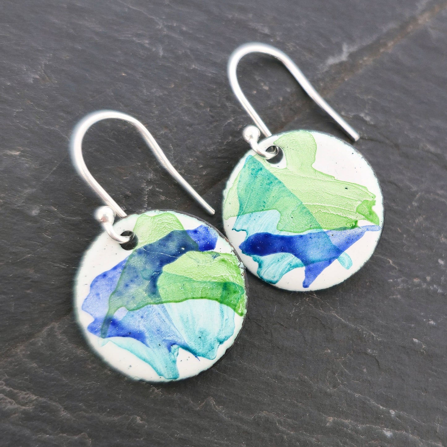 Round enamel drop earrings with splashes of dark blue and green on a white background with silver ear hooks. Handmade. Mijoux Jewellery.