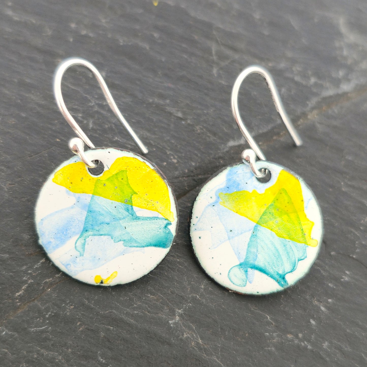Round enamel drop earrings with splashes of blue, green and yellow on a white background. On silver ear hooks. Mijoux Jewellery.