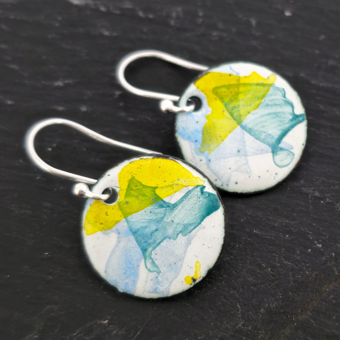 Round enamel drop earrings with splashes of blue, green and yellow on a white background. On silver ear hooks. Mijoux Jewellery.