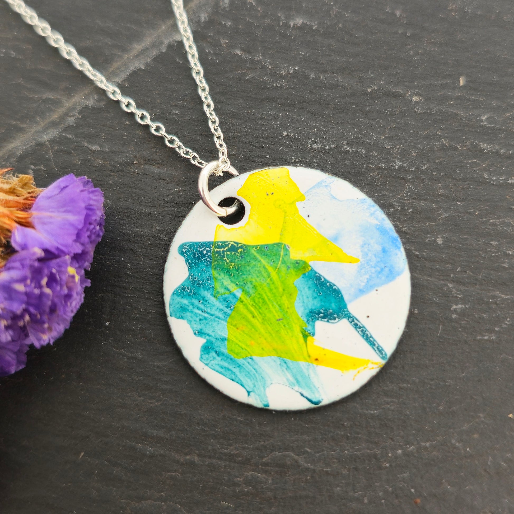 A round enamel pendant with splashes of yellow, green and blue. Suspended on a silver chain. Shown on a black background with purple flowers. Mijoux Jewellery.