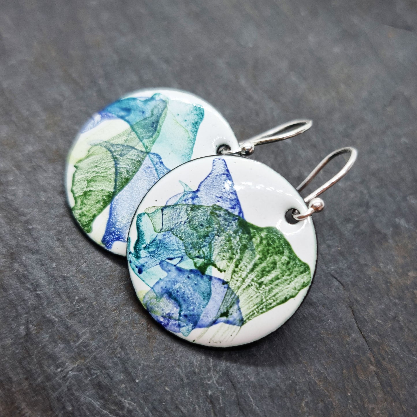 Round enamel drop earrings with splashes of dark blue and green on a white background.