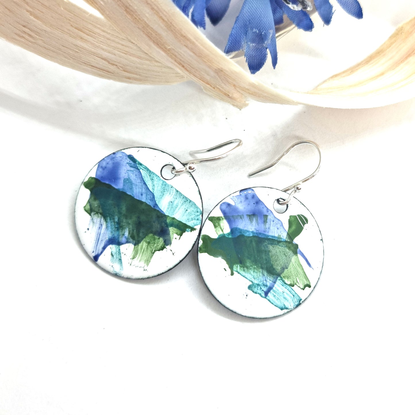 Round enamel drop earrings with splashes of dark blue and green on a white background with flower and wood shavings