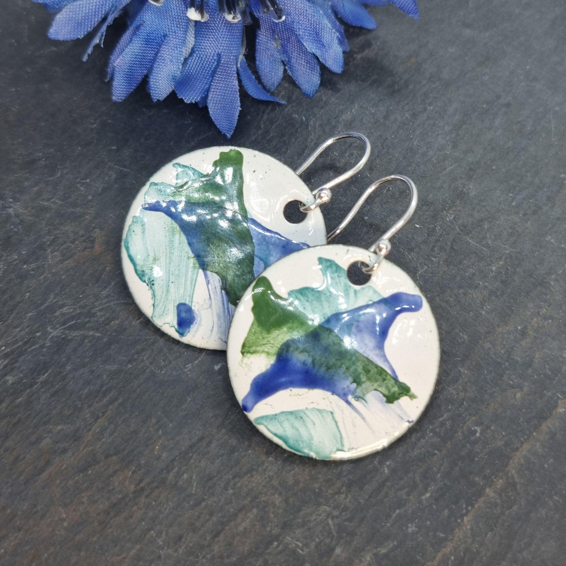 Round enamel drop earrings with splashes of dark blue and green on a white background with flower