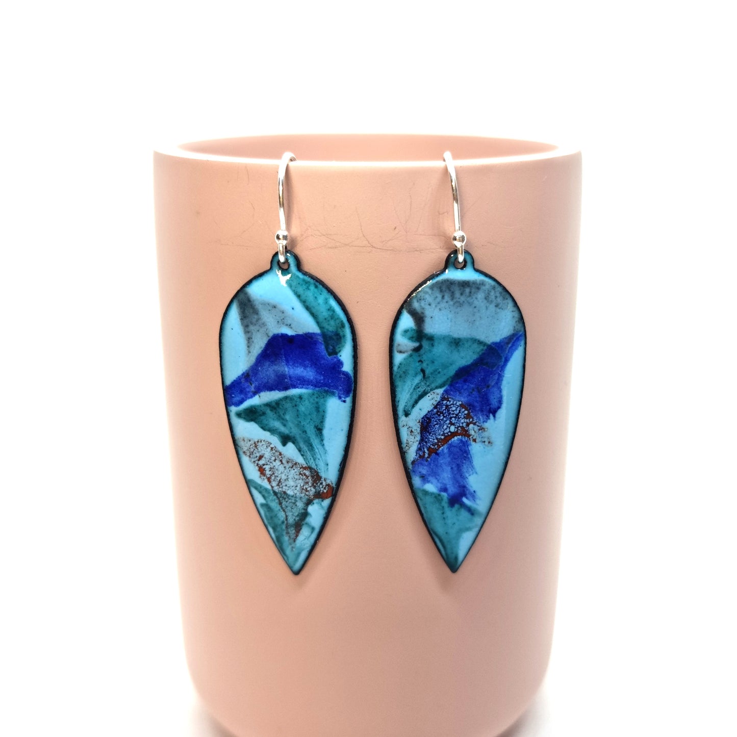 Large teardrop shaped enamel drop earrings with splashes of dark blue, red, green and grey on a light blue background