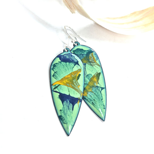 Large teardrop-shaped enamel drop earrings with splashes of dark blue, green and yellow on a light green background - with wood shavings