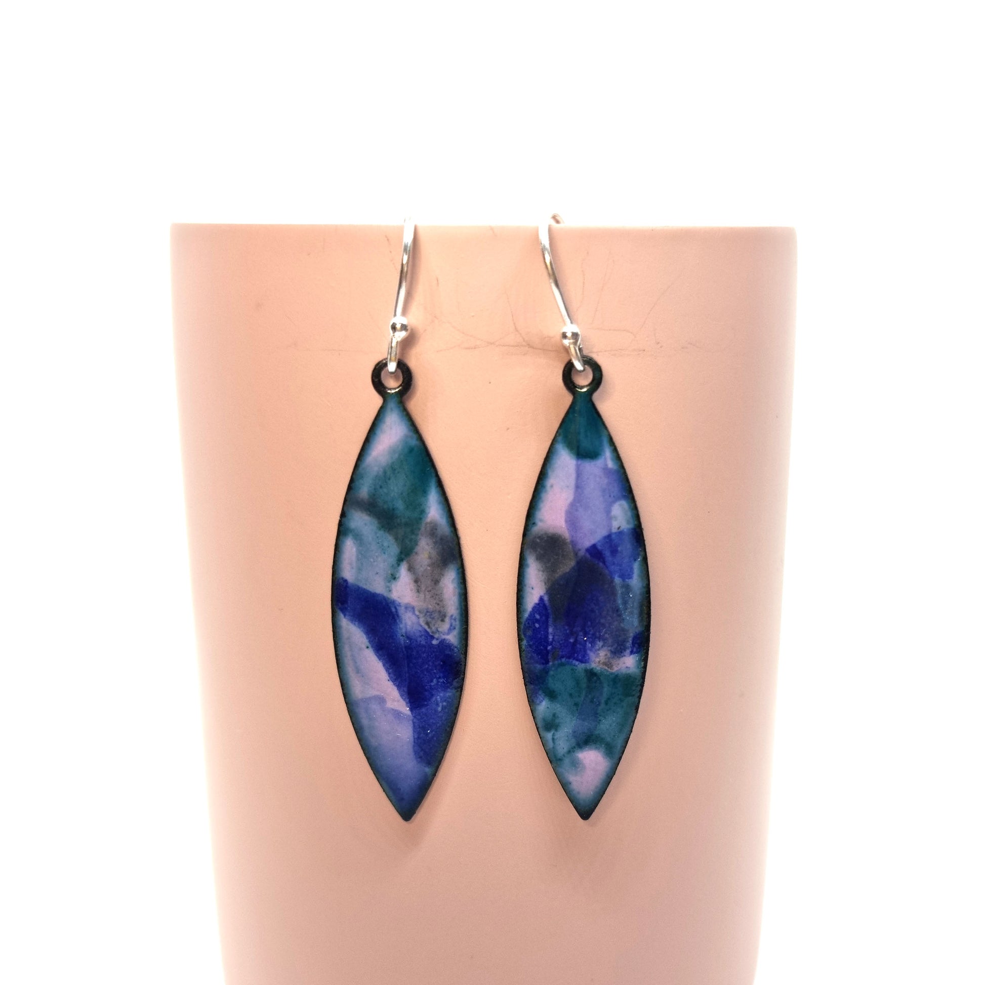 Boat-shaped enamel drop earrings with splashes of blue, green and grey on a light purple background