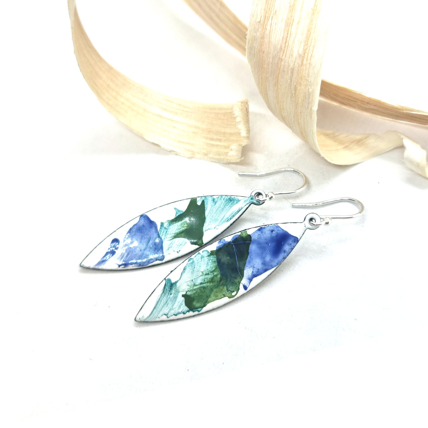 Navette shaped enamel drop earrings with splashes of green and blue on a white background with wood shavings