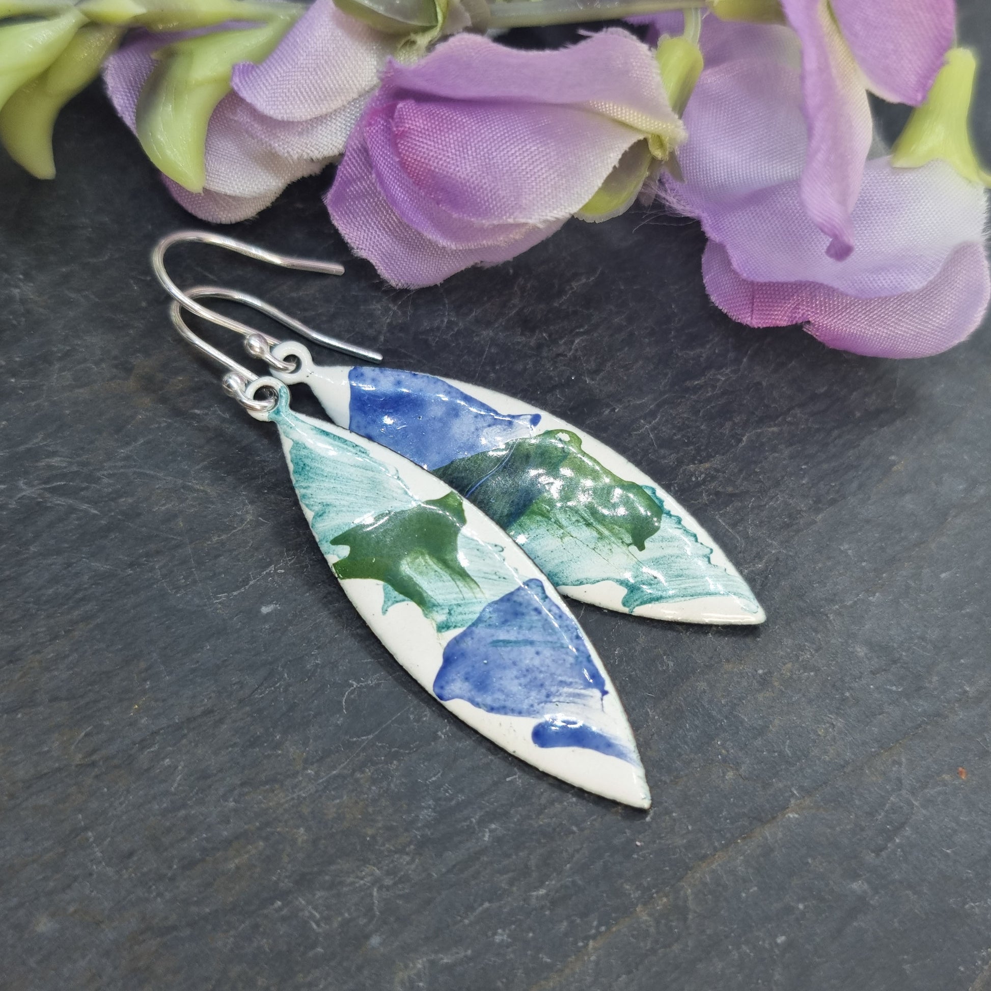 Navette shaped enamel drop earrings with splashes of green and blue on a white background with flowers