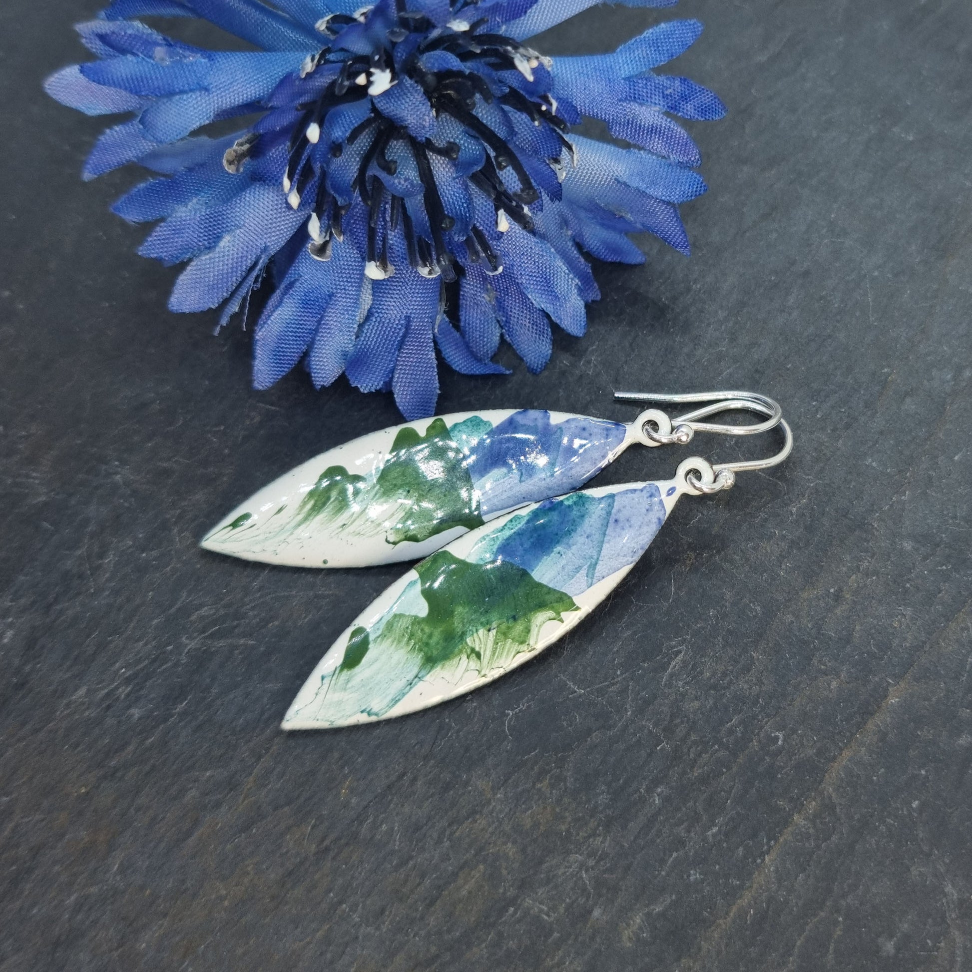 Navette shaped enamel drop earrings with splashes of green and blue on a white background with flower