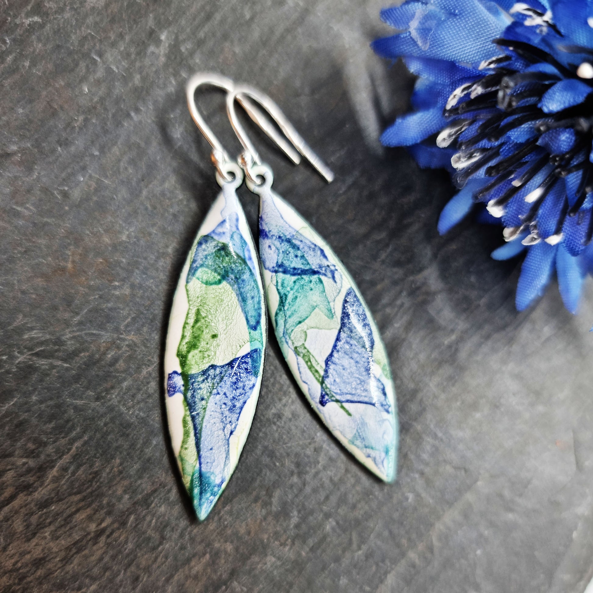 Navette shaped enamel drop earrings with splashes of green and blue on a white background with flower