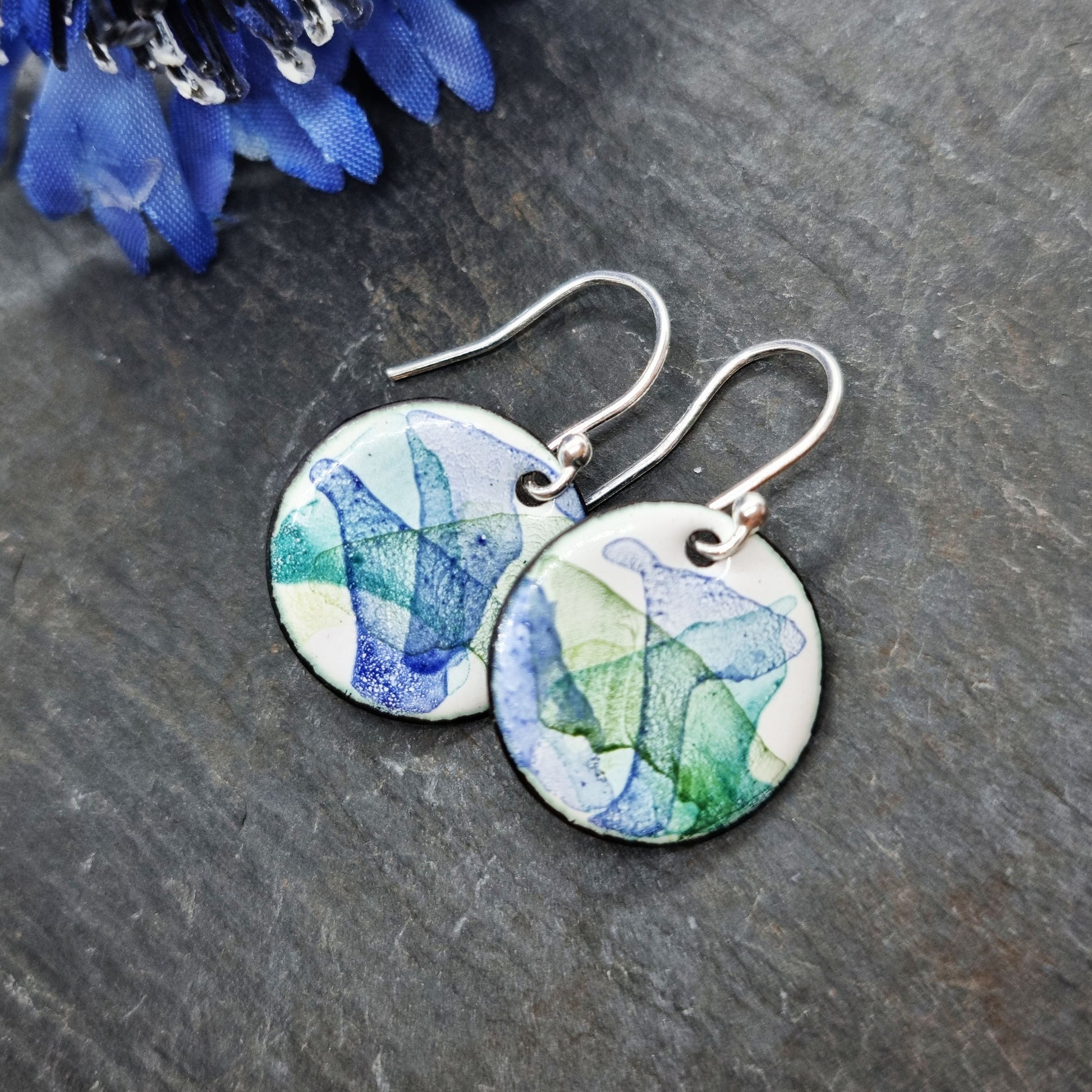 Round enamel drop earrings with splashes of dark blue and green on a white background - with flower