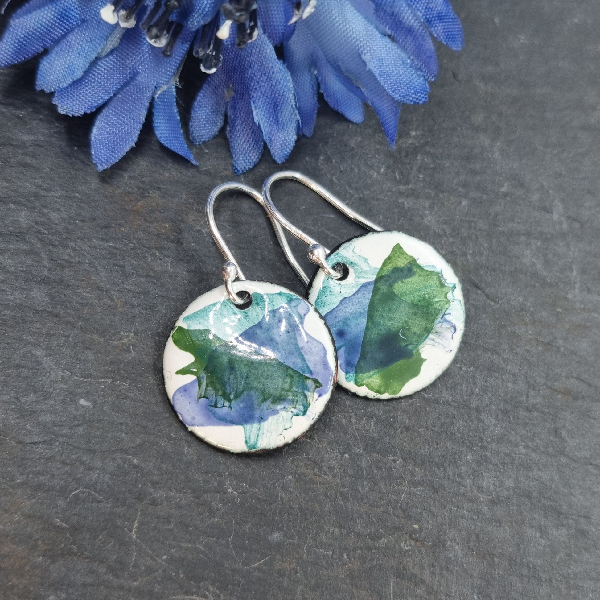 Round enamel drop earrings with splashes of dark blue and green on a white background with flower