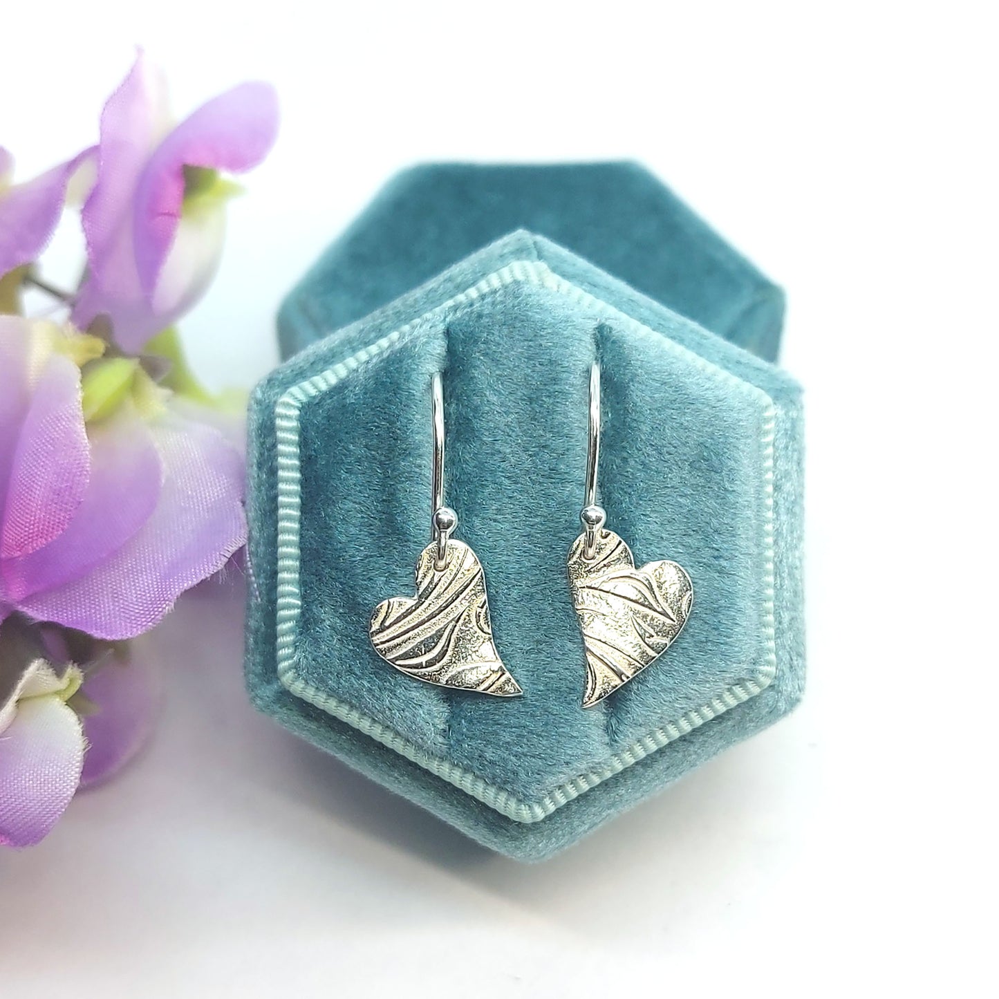 Silver heart drop earrings featuring an asymmetrical heart with a leaf and vine pattern suspended from silver ear hooks. Shown in a jewellery box with flowers.