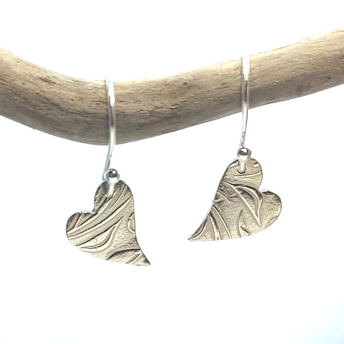 Silver heart drop earrings featuring an asymmetrical heart with a leaf and vine pattern suspended from silver ear hooks. Shown suspended from a twig.